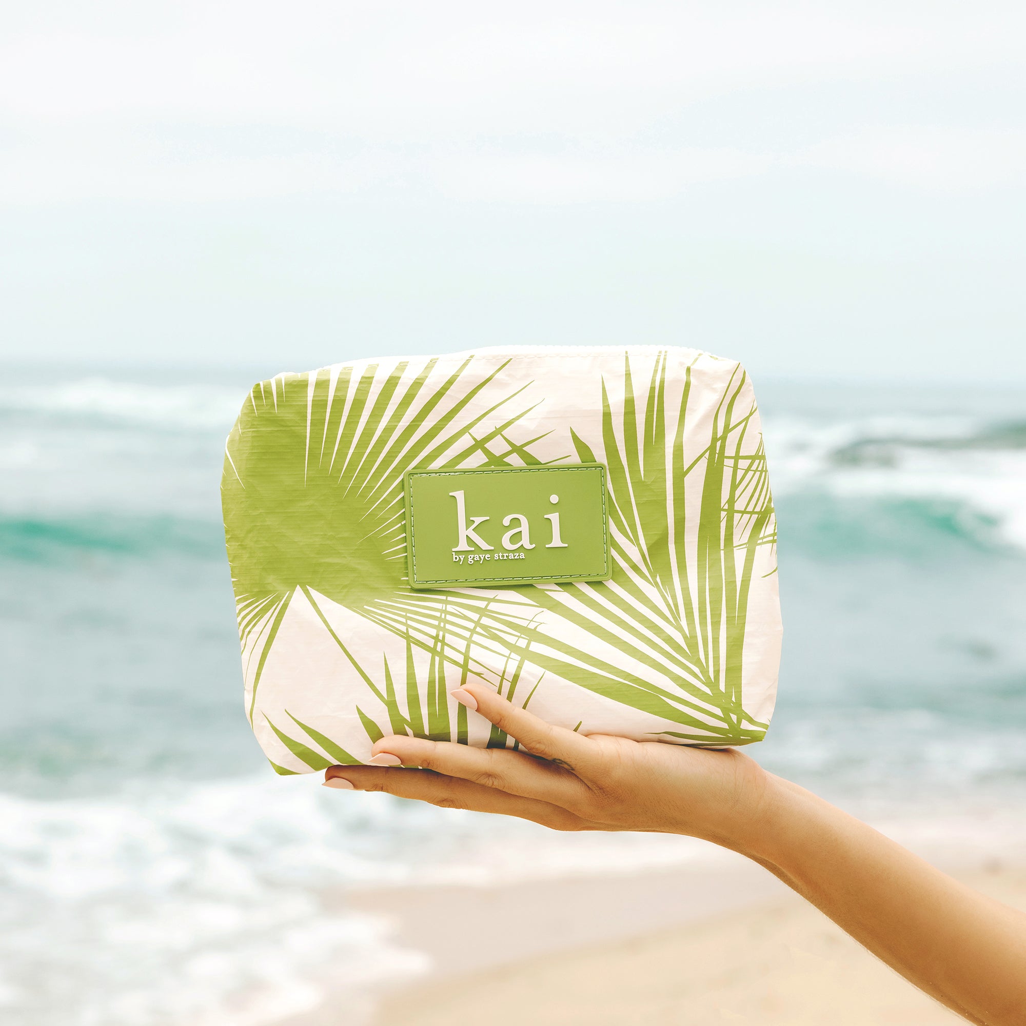 hand holding a green and white palm leaf patterned aloha pouch with kai brand logo against a beach background.