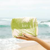 hand holding a green and white palm leaf patterned aloha pouch with kai brand logo against a beach background.